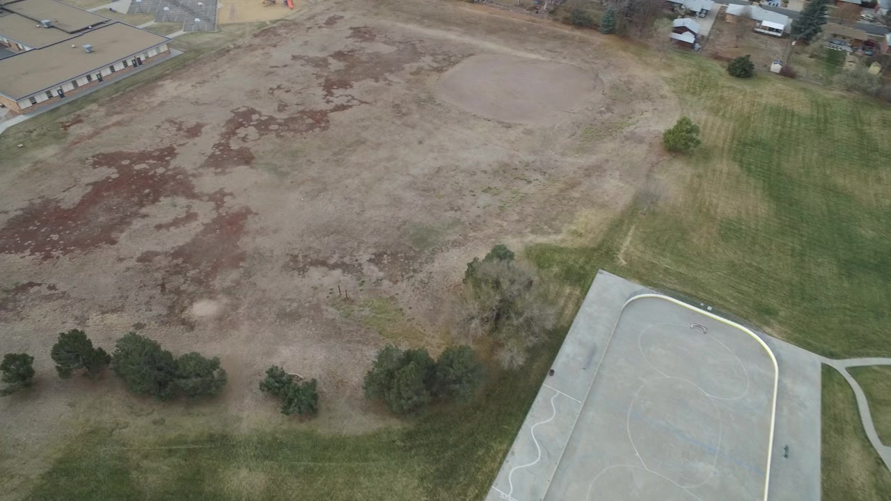 Aerial view over patchy empty land beside a paved recreation area and low buildings.