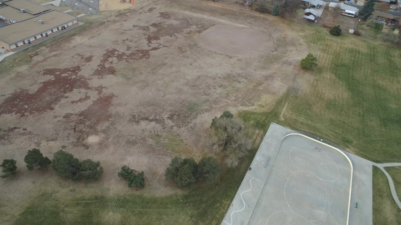 Aerial view in Westminster, Colorado with the empty lot centered, a low tan building at the top, green grass bands, scattered trees, and no visible people.