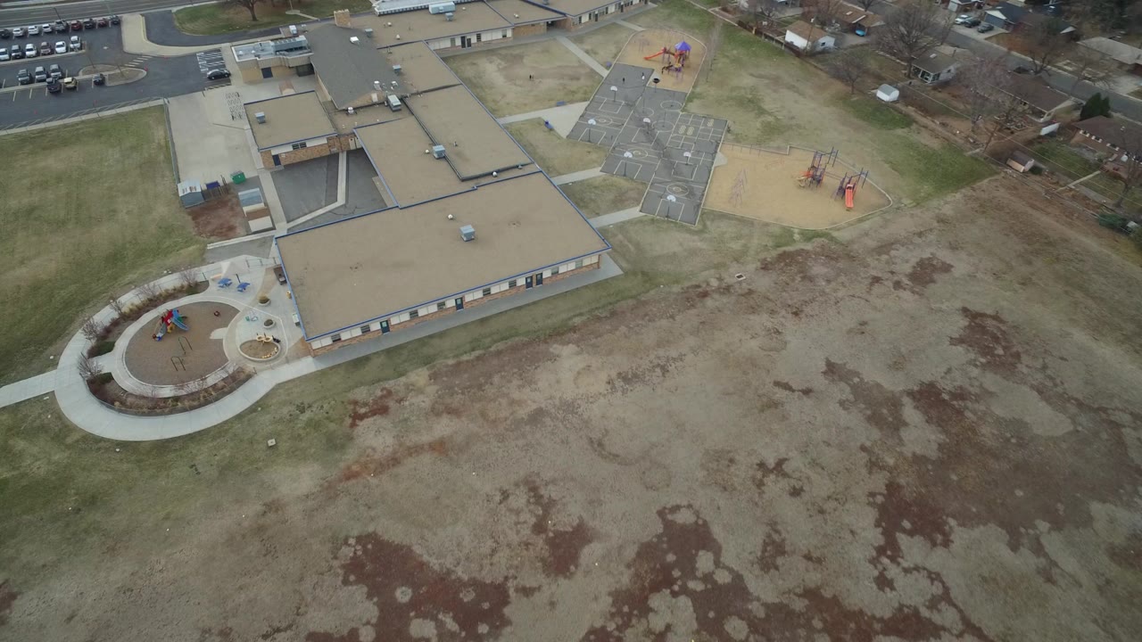 Aerial view in Westminster, Colorado showing a large tan-roofed building, curved sidewalk, playground areas, parking lot, nearby homes, and no visible people.