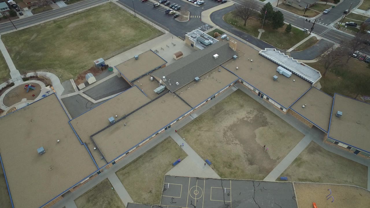 Overhead aerial frame in Westminster, Colorado showing a flat-roofed campus, patchy grass areas, roads, sidewalks, and basketball courts with a few tiny people visible.