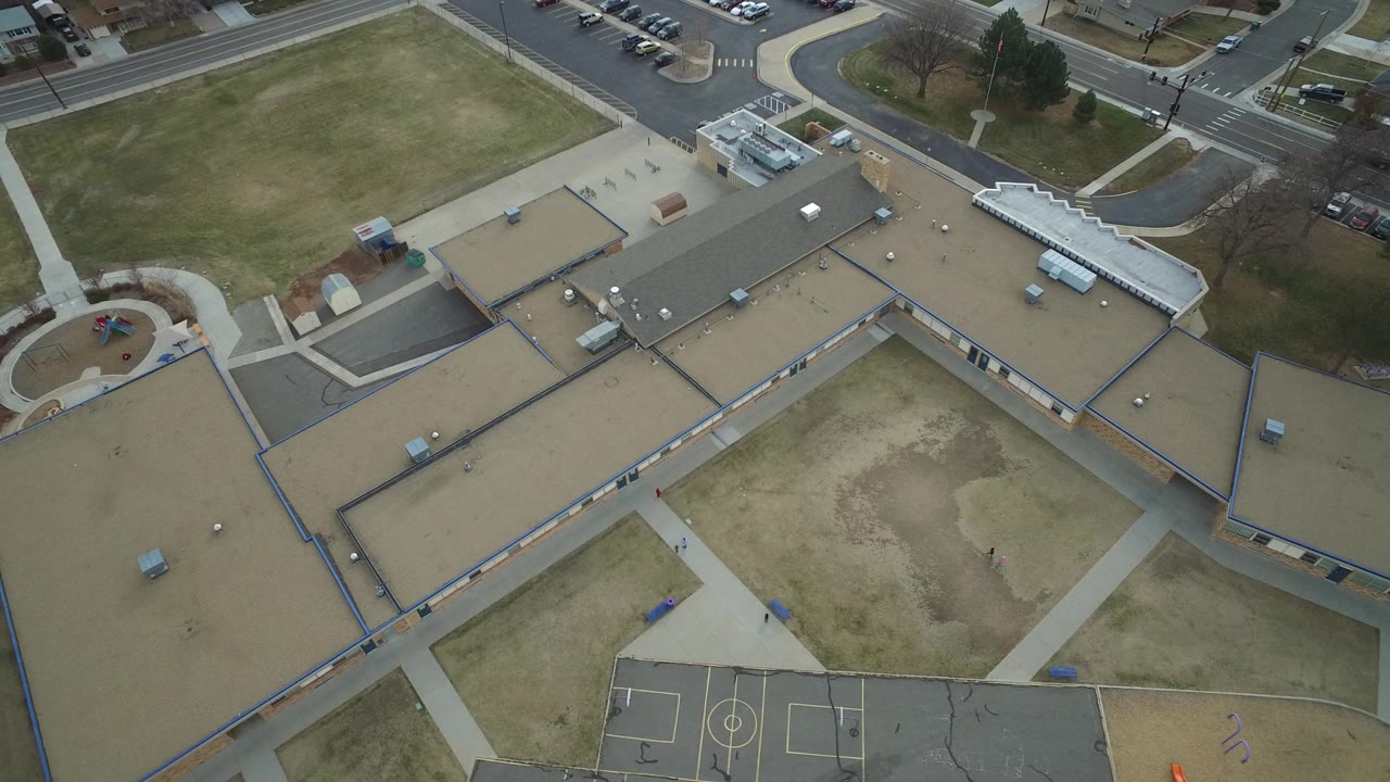 Westminster, Colorado aerial frame looking down on tan and gray school rooftops, open grass, intersecting sidewalks, parked cars, and a court area along the lower edge.