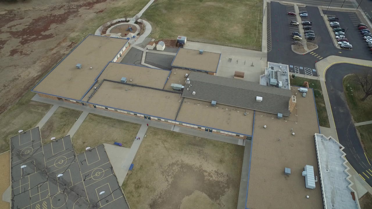 Overhead aerial frame in Westminster, Colorado showing tan flat roofs, dark sports courts at lower left, a parking lot with cars at upper right, winter-brown grass, and no visible people.