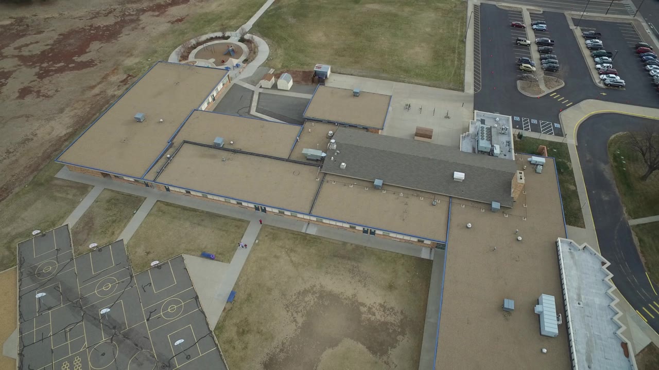 Aerial view in Westminster, Colorado with the building roofs centered, sports courts in the lower left corner, a curved paved drive on the right, a parking lot beyond it, and no visible people.