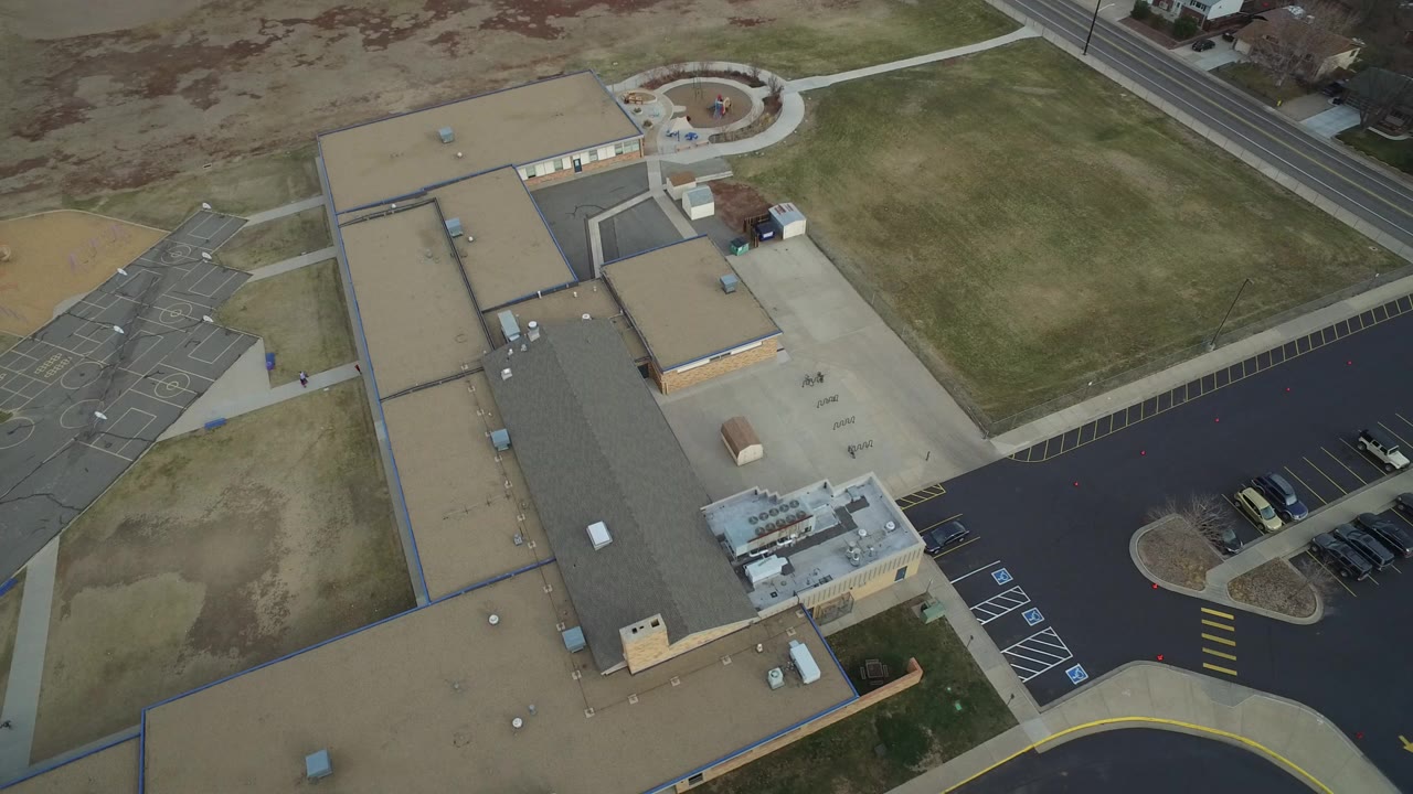 High overhead frame of a Westminster, Colorado building complex with tan roofs, blacktop parking, sidewalks, lawns, and no visible people at the start of the clip.