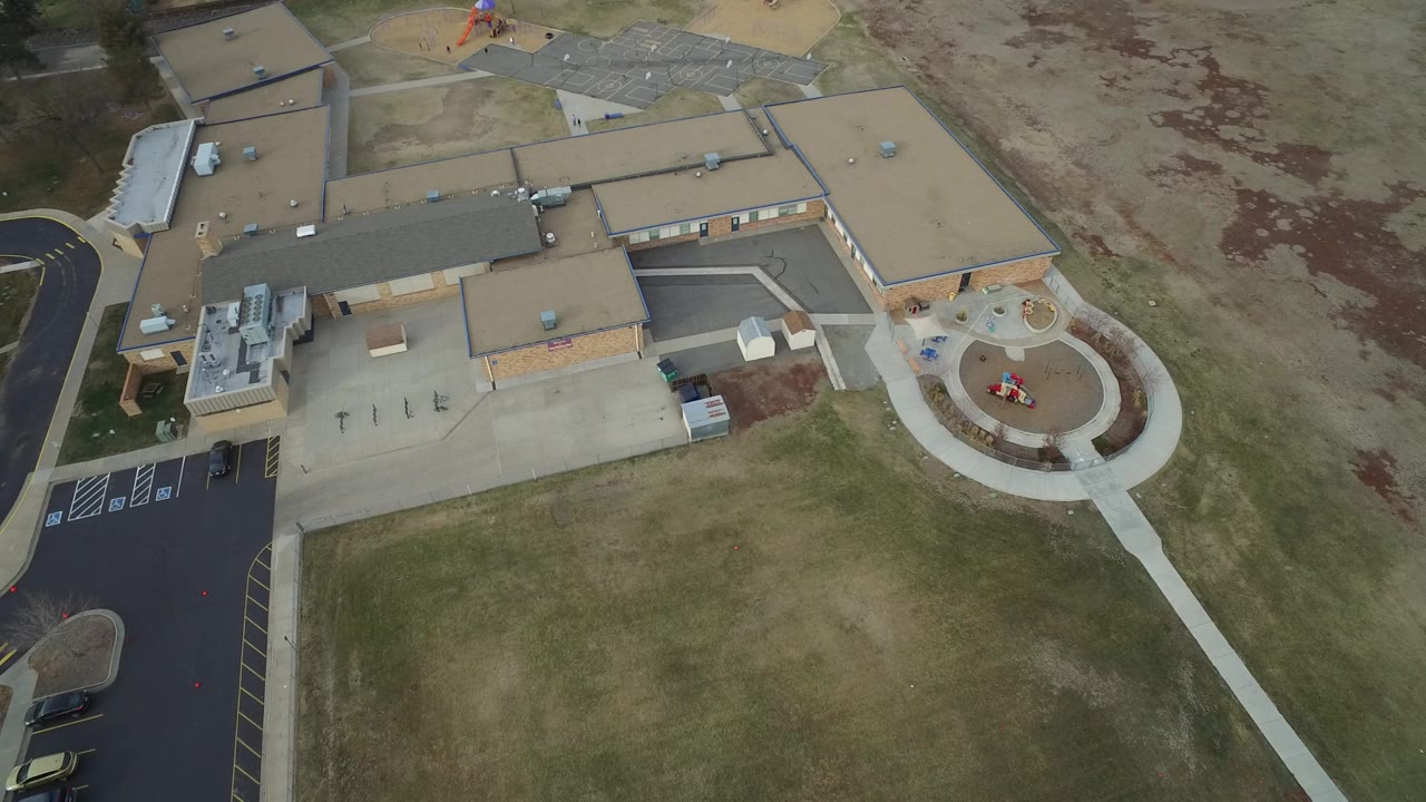 Overhead view in Westminster, Colorado showing a tan-roofed suburban building, blacktop parking at left, dry greenish grass below, and a circular play area at right with no clearly visible people.