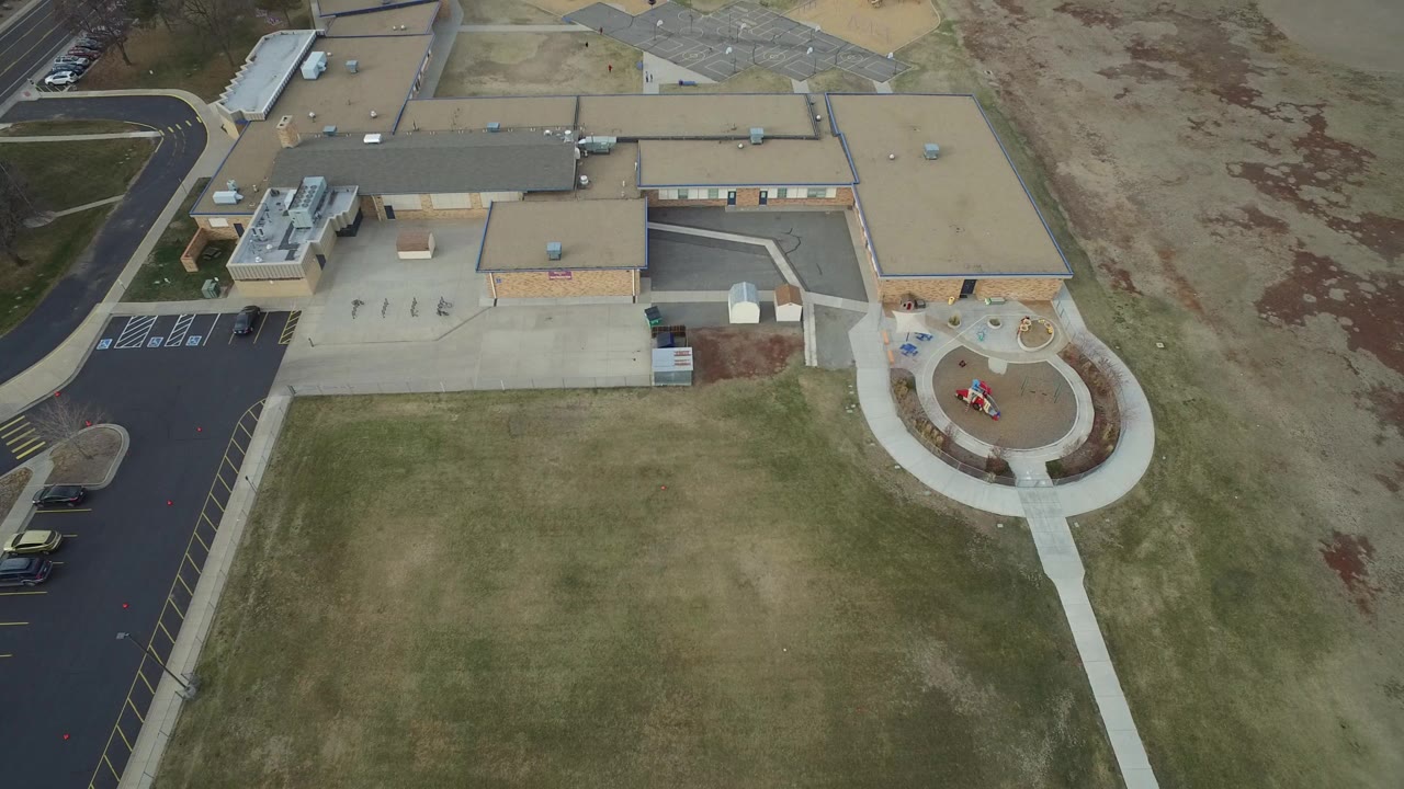 Overhead view of a large low-rise building, parking lot, sidewalks, grass, and playground area.