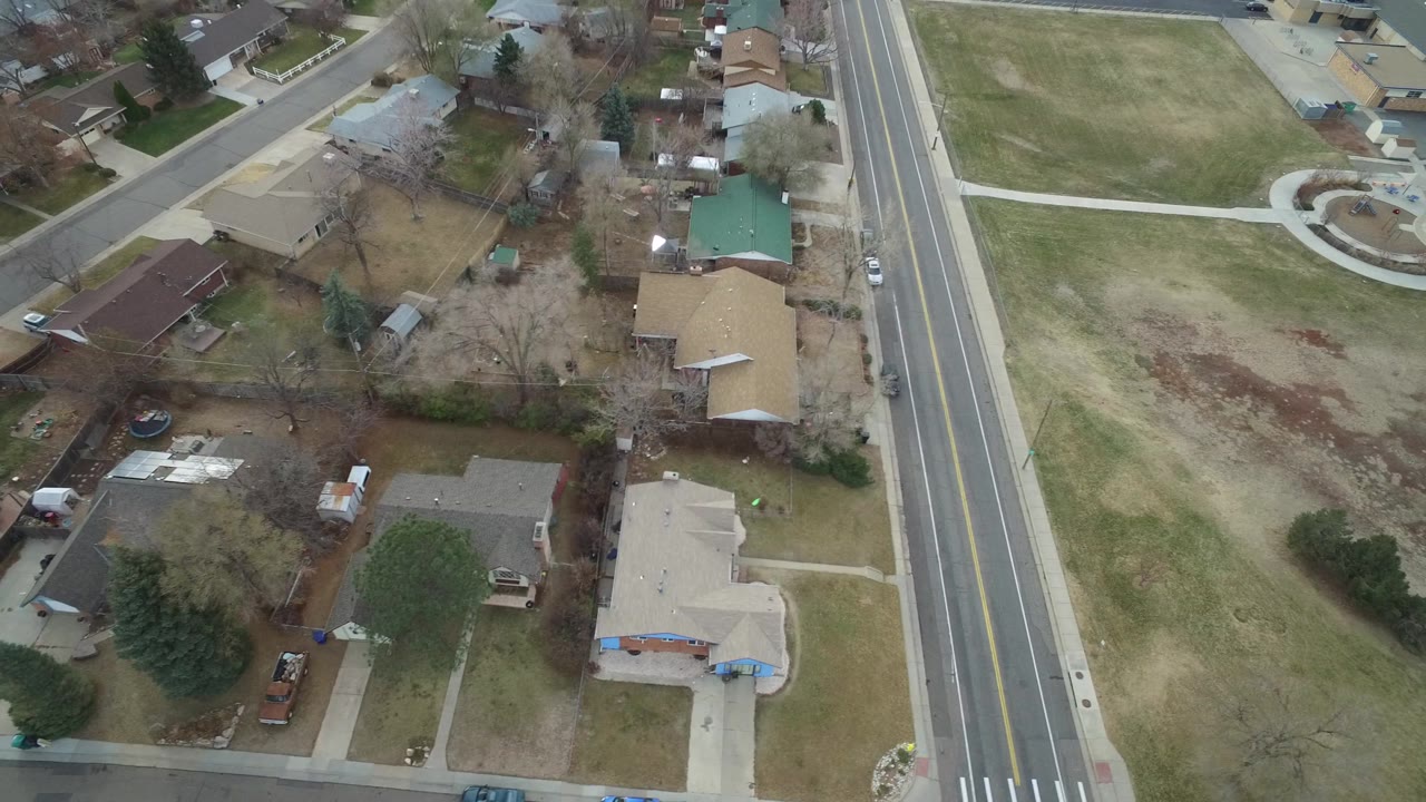 Overhead Westminster, Colorado neighborhood view with rooftops, fenced yards, bare trees, parked cars, and the main road running along the right side, with no visible people.
