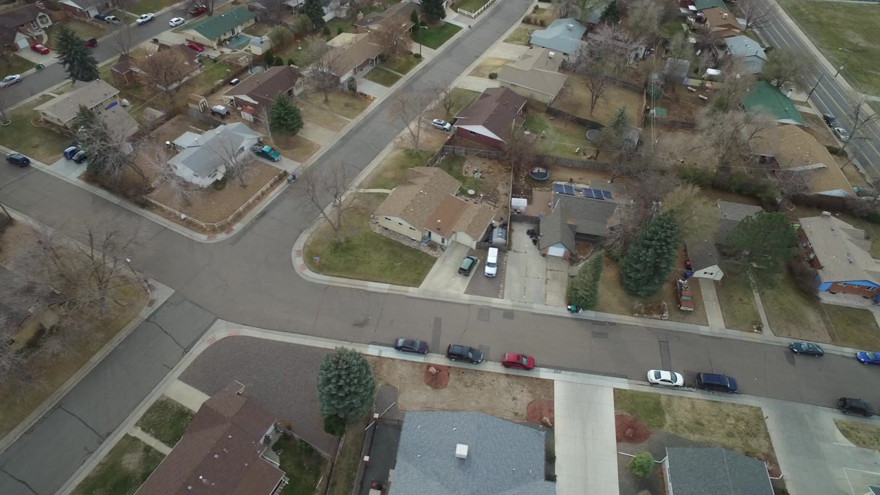 Bird-view frame in Westminster, Colorado showing rooftops, driveways, small lawns, parked vehicles, and leafless trees across a residential block with no visible people.