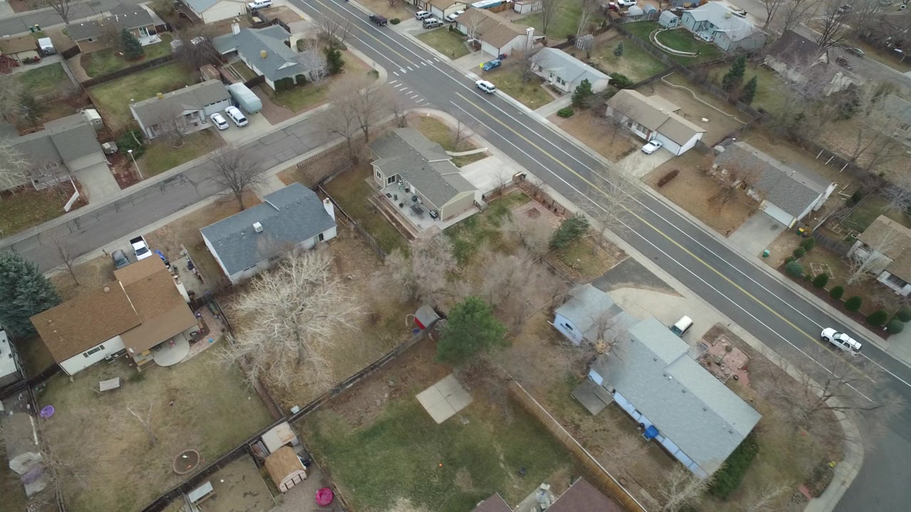 High aerial frame over Westminster, Colorado showing single-family homes, fenced yards, parked cars, leafless trees, and a diagonal multi-lane road with no visible people.