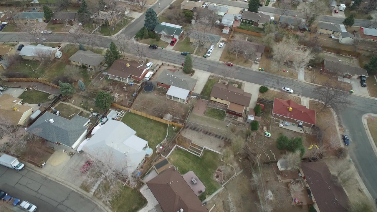High-angle Westminster, Colorado suburb frame showing rooftops, a curved residential street, fences, lawns, parked vehicles, and no visible people.