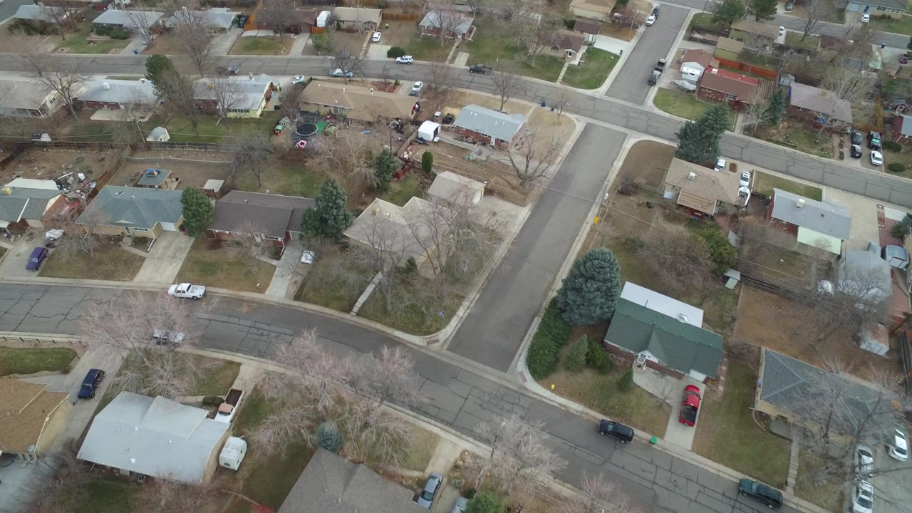 High aerial view over Westminster, Colorado residential streets, showing detached homes, driveways, brown lawns, parked vehicles, and no visible people at the start of the clip.