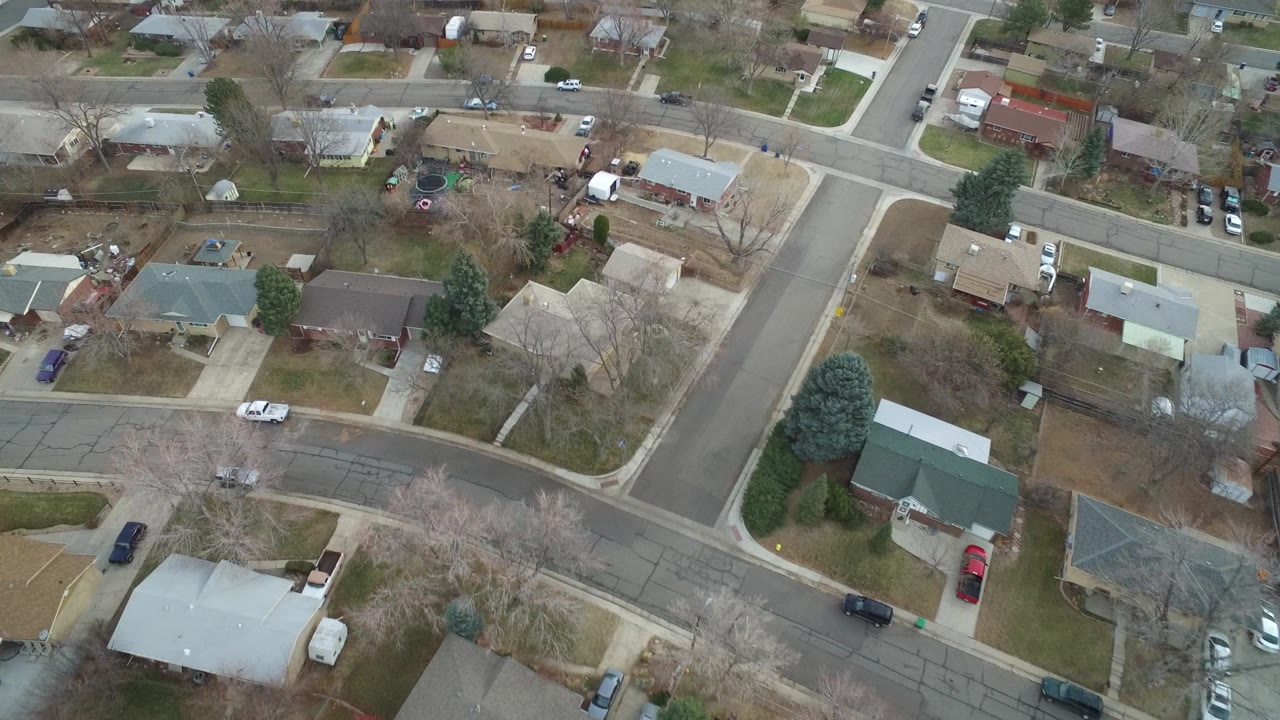 Overhead Westminster, Colorado neighborhood view with a cross street, single-family roofs, small yards, parked cars, and no visible people one second into the clip.