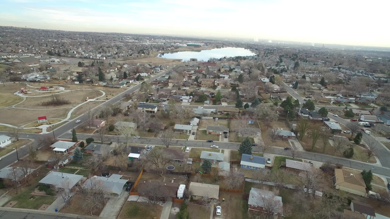 Wide aerial frame over Westminster, Colorado homes, intersecting roads, open grass, bare trees, and a distant lake under a pale sky, with no visible people.