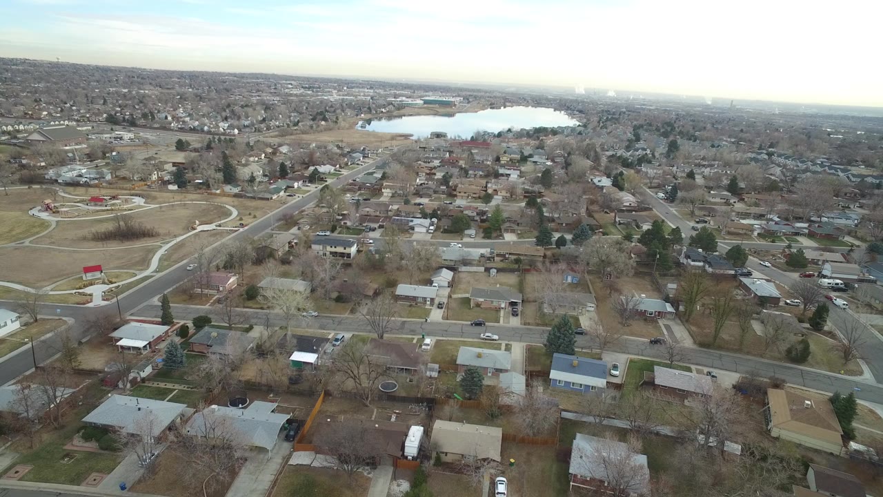 Drone-style view of homes, streets, open fields, and a distant lake.