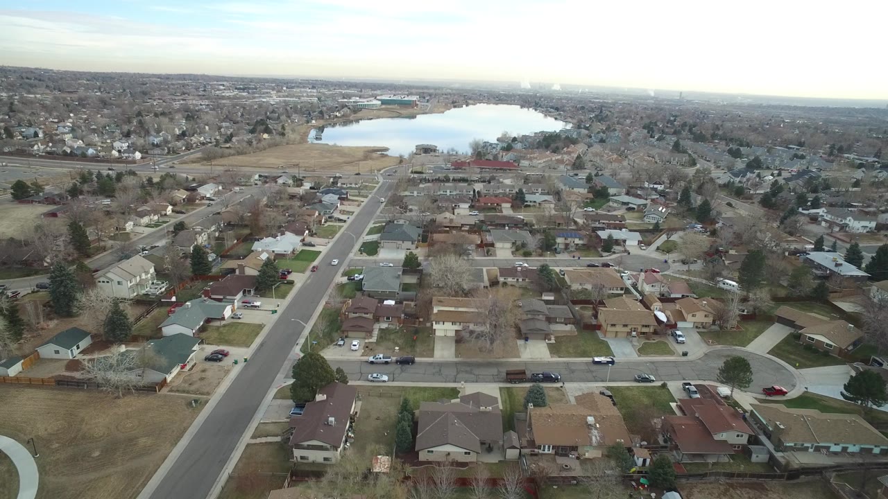 Aerial frame over Westminster, Colorado residential blocks with rooftops, parked cars, bare trees, and the lake centered near the horizon, with no visible people.