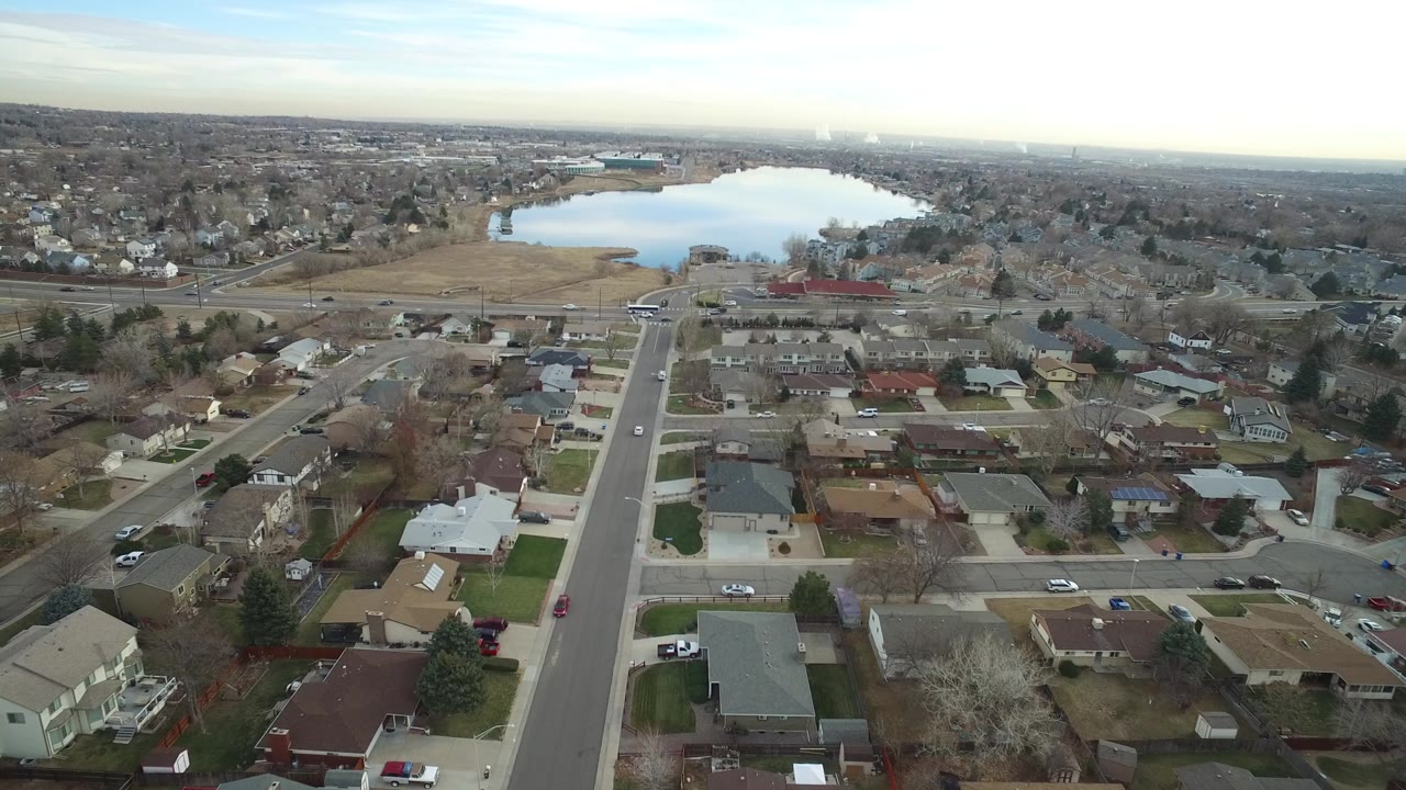 High drone view across Westminster, Colorado residential blocks, with cars on neighborhood roads, bare trees around homes, and the lake centered near the horizon.