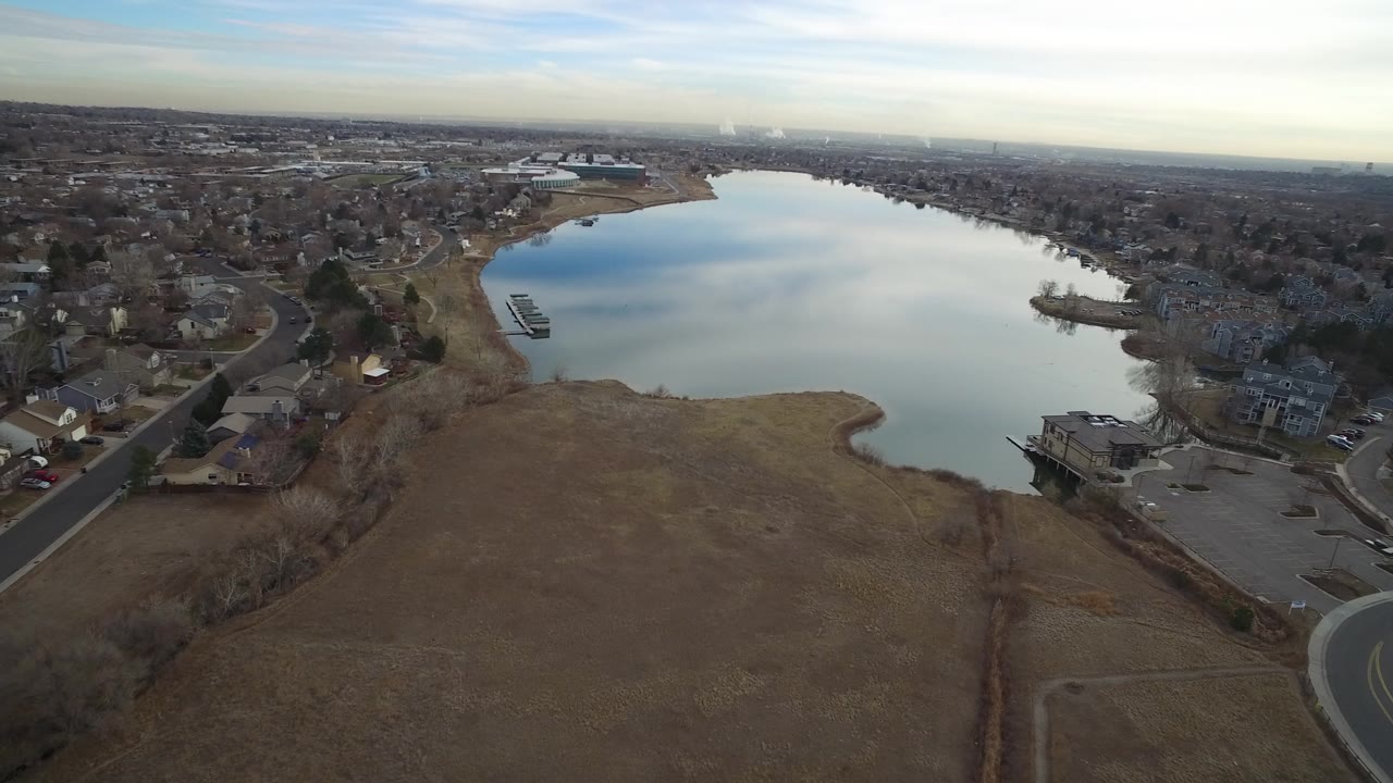 Aerial view over a lake with suburban homes and brown shoreline grass.