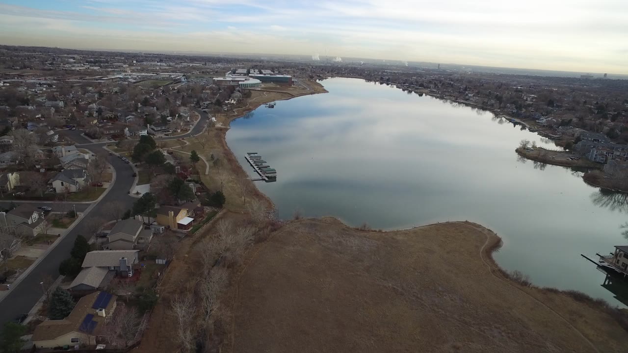 High aerial view in Westminster, Colorado looking across a wide lake, curved residential streets, shoreline grass, small docks, distant development, and no visible people.