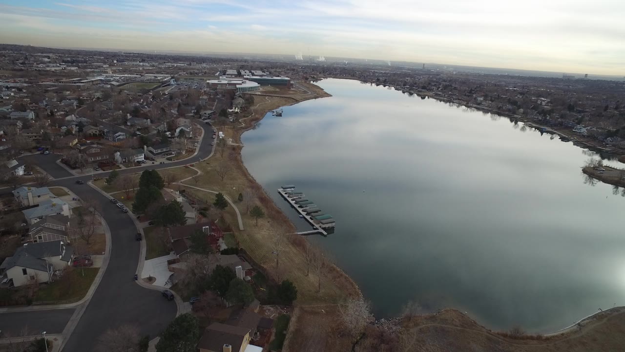Aerial view in Westminster, Colorado looking across a broad lake bordered by houses, curved streets, dry shoreline grass, docks, and distant low buildings, with no visible people.