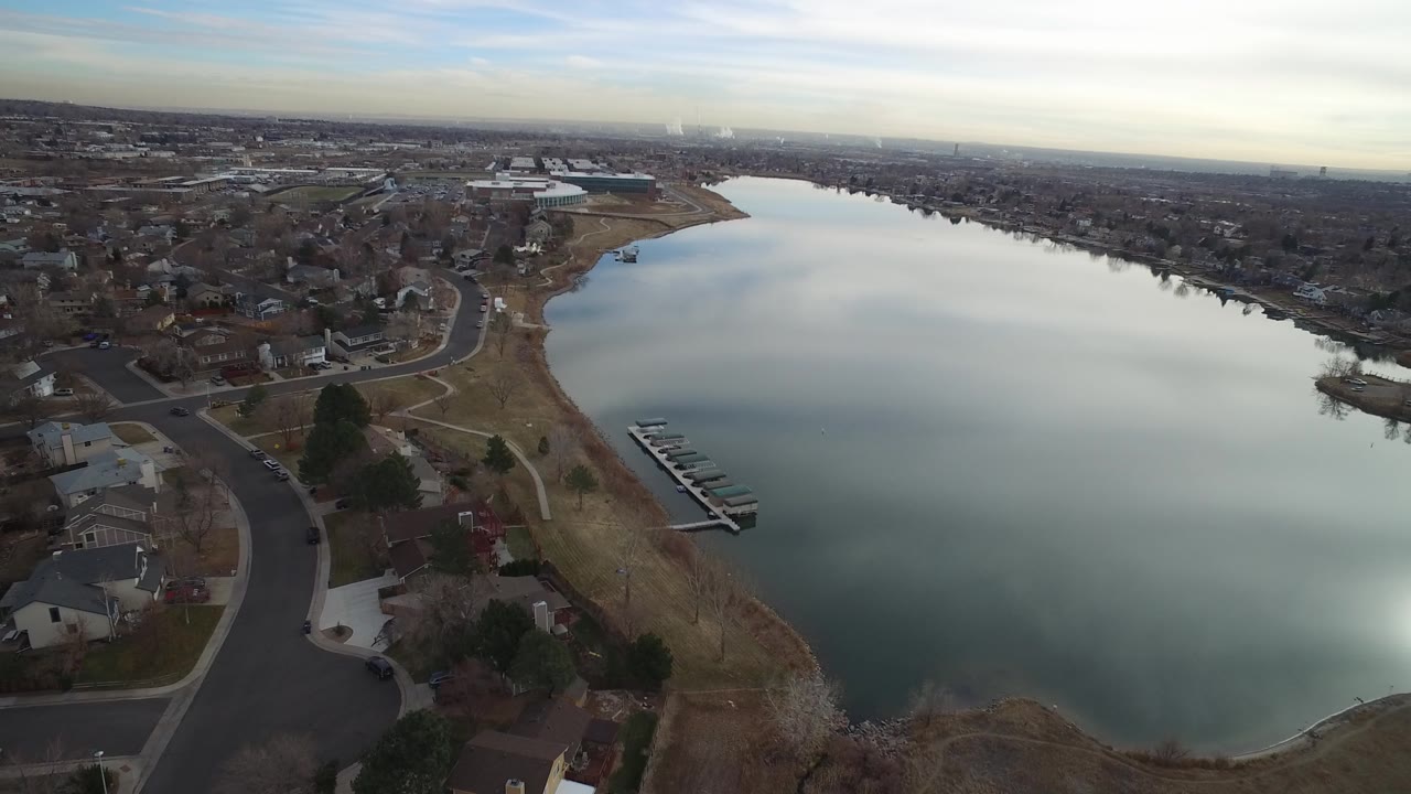 A second aerial view in Westminster, Colorado shows the lake surface occupying more of the frame, with neighborhood homes, a curved road, shoreline paths, and a small dock cluster visible below, with no visible people.