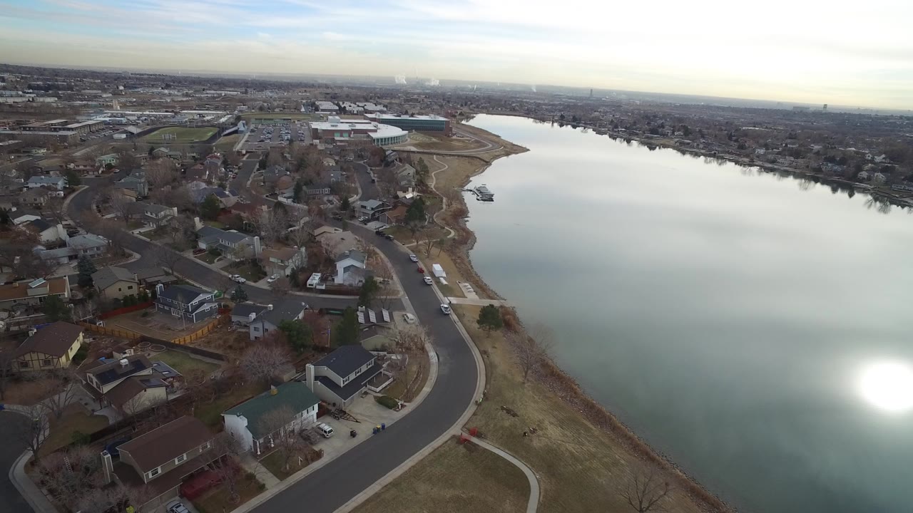 Opening aerial frame over Westminster, Colorado, showing calm water with a bright sun reflection, shoreline houses, curving streets, bare trees, and no visible people.