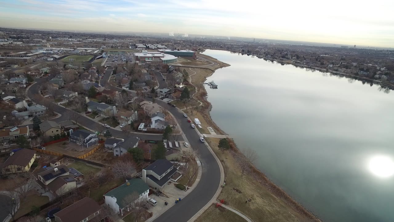 High aerial over a suburban lakeshore with homes, streets, and bright glare on the water.
