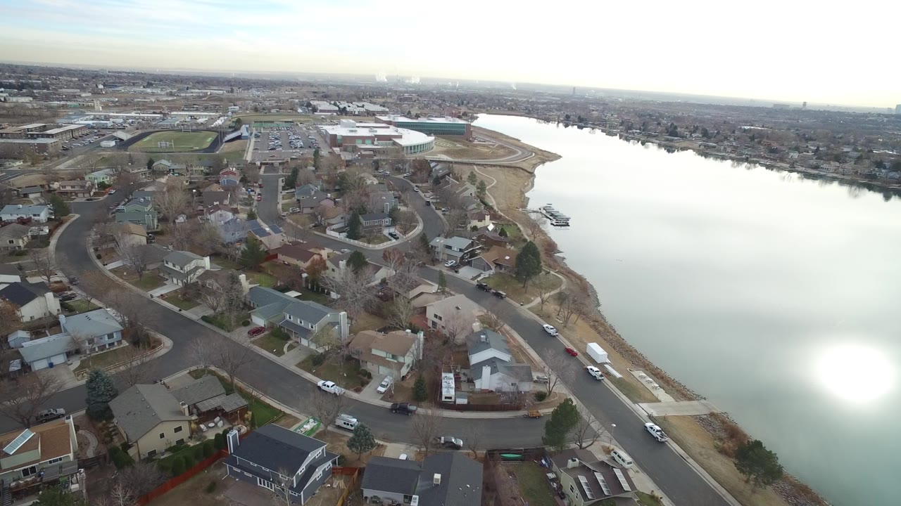 Drone-like aerial frame over Westminster, Colorado showing a residential street curve, rooftops, parked vehicles, a reflective river, and no visible people.
