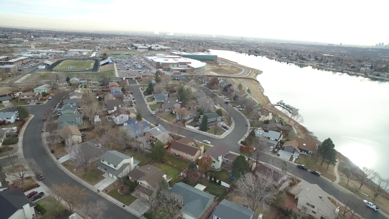 Second aerial frame in Westminster, Colorado showing rooftops and winding roads in the foreground, the lake along the right, a parking area and sports field ahead, and no visible people.
