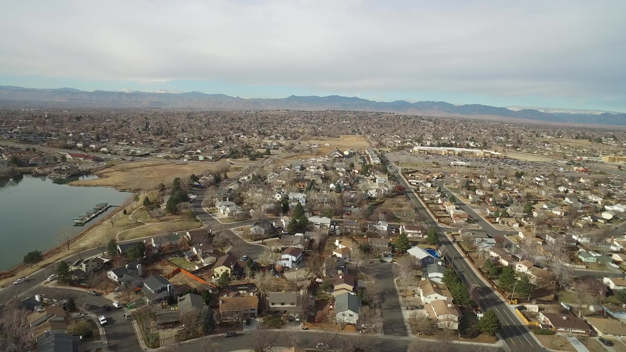 Aerial view over Westminster, Colorado showing a lake at left, suburban homes and streets below, distant mountains, cloudy sky, and no visible people.