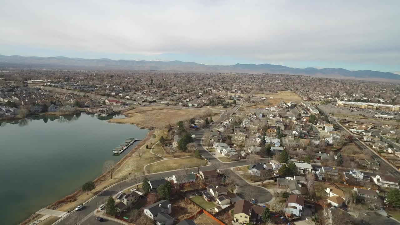 Aerial view over Westminster, Colorado showing a large lake at left, nearby homes and roads at right, distant mountains on the horizon, and no visible people.