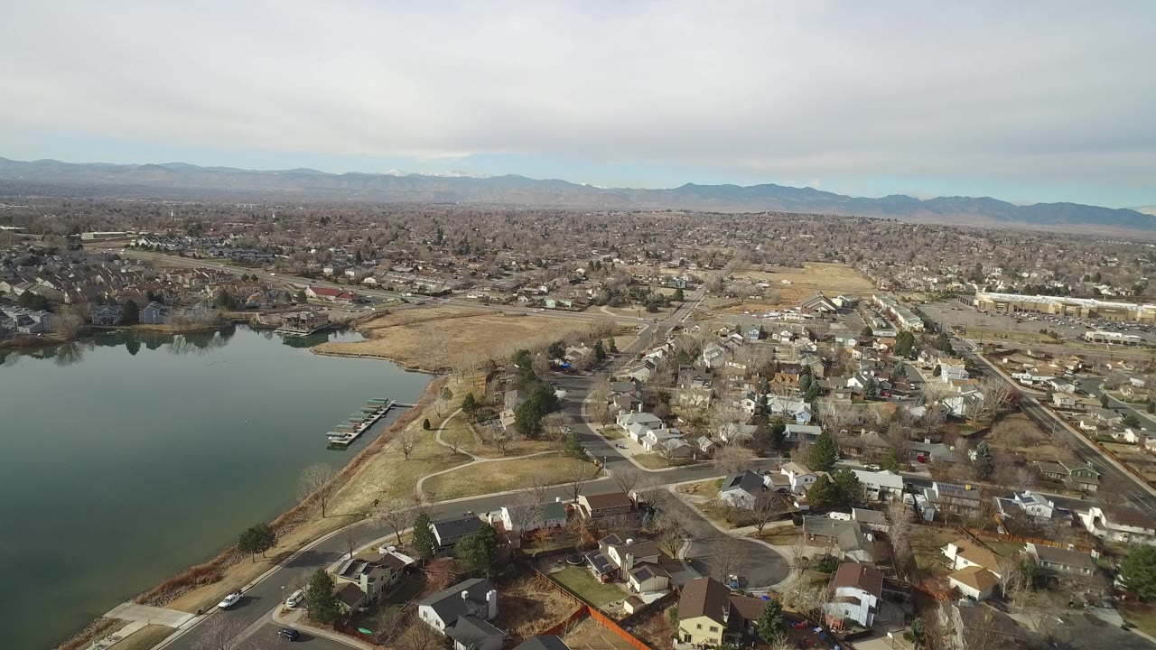 Wide aerial frame in Westminster, Colorado with the lake occupying the left foreground, a dock near the shoreline, clustered houses to the right, and no visible people.