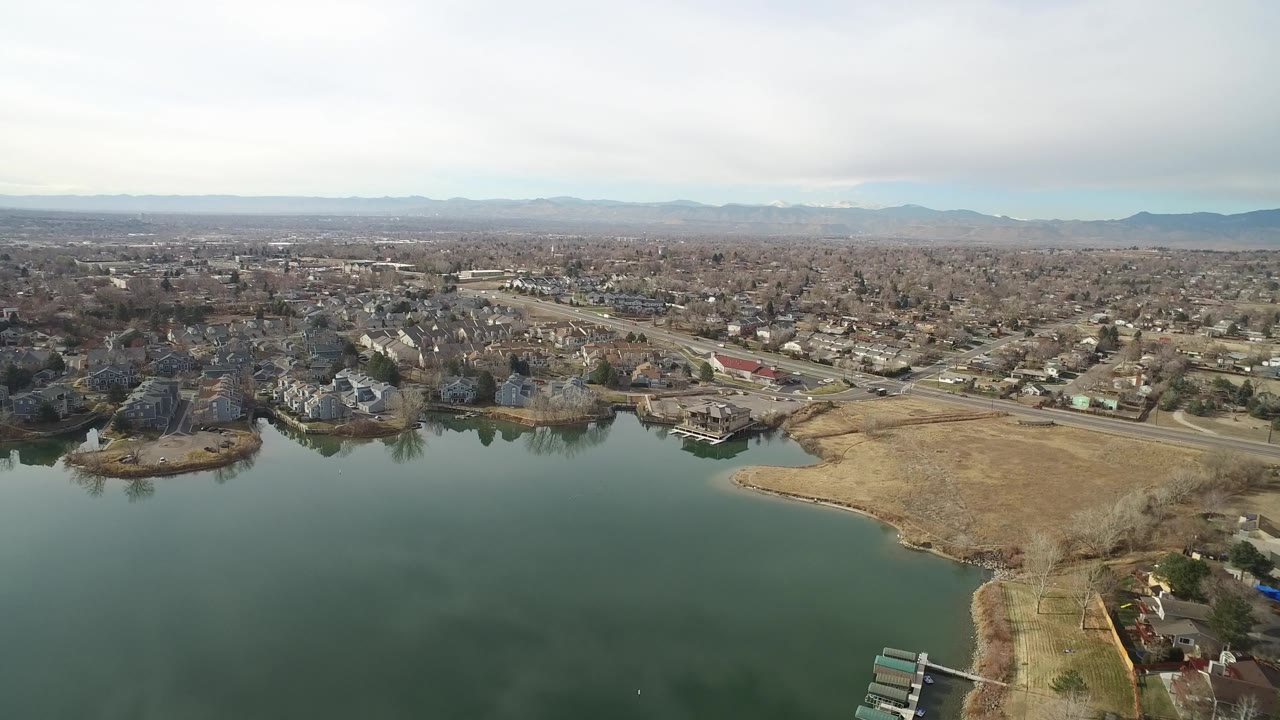 Aerial view in Westminster, Colorado, looking past lake water and a tan shoreline toward clusters of houses, a road corridor, distant low-rise buildings, and no visible people.