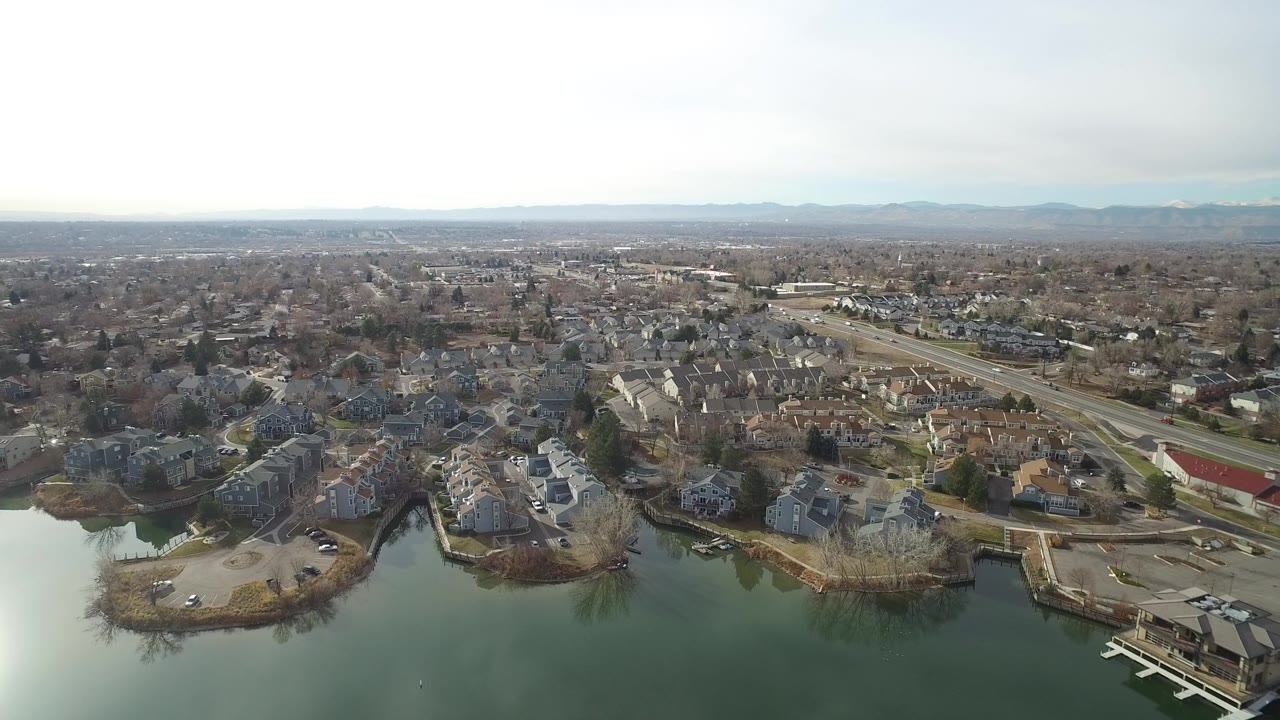 Wide aerial view over Westminster, Colorado homes beside a greenish lake, with a multi-lane road, bare trees, hazy horizon, and no visible people.