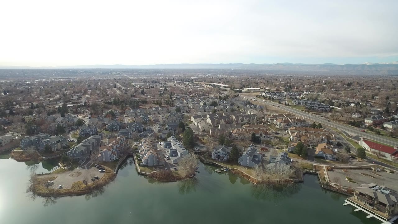 Aerial frame over Westminster, Colorado with lake water in the foreground, residential buildings along the shore, road traffic beyond, and no visible people.