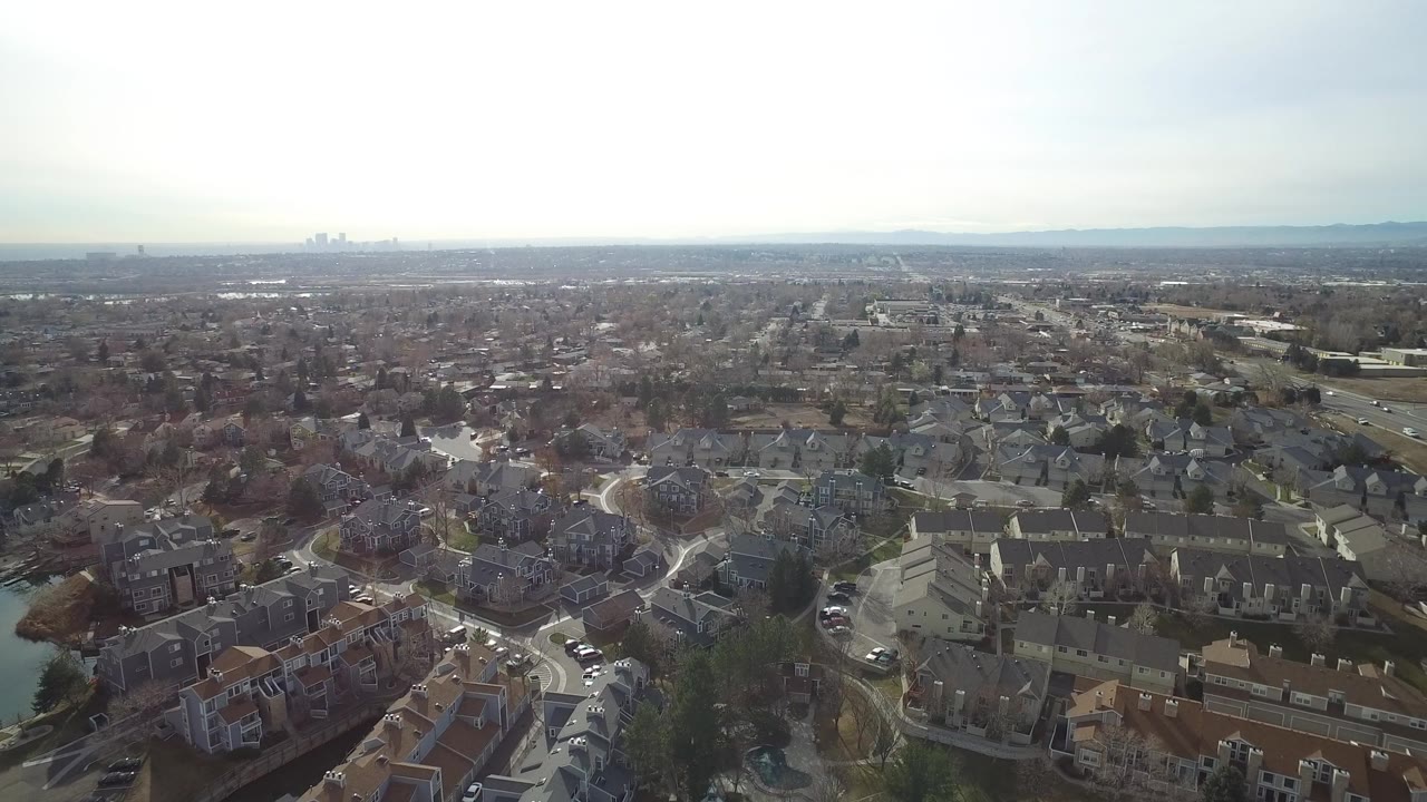 Aerial view over dense suburban homes and curving streets under a bright hazy sky.