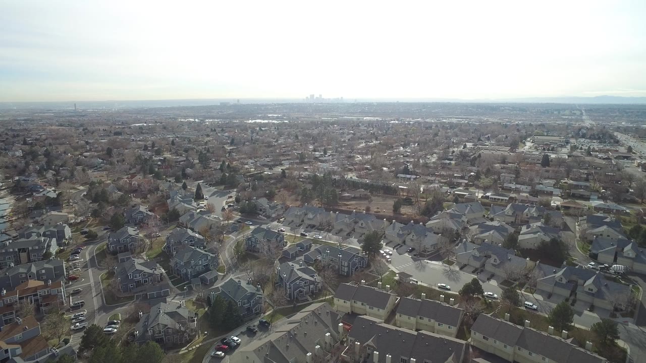 High aerial view over Westminster, Colorado rooftops, curving residential streets, bare trees, parked cars, and a hazy horizon with no visible people.
