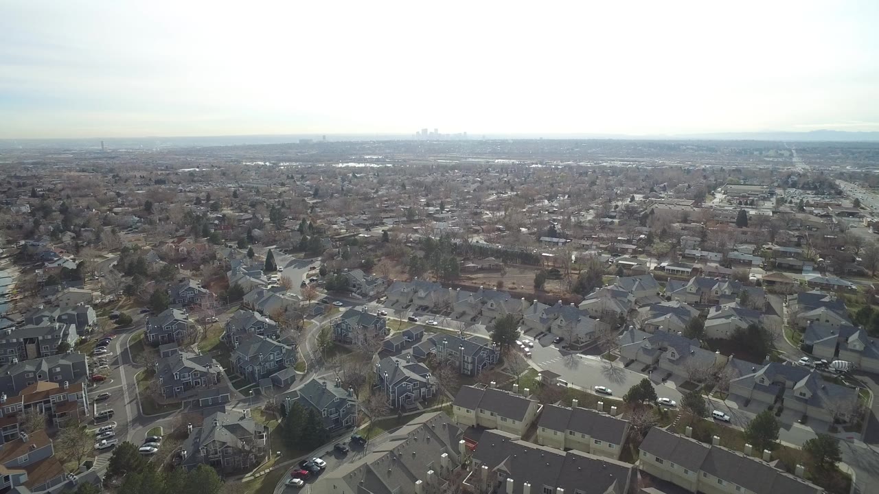 Aerial frame in Westminster, Colorado showing dense residential blocks, gray rooftops, scattered parked cars, winter trees, and distant city buildings with no visible people.