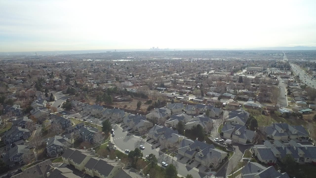 Aerial frame over Westminster, Colorado showing gray rooftops in the foreground, residential blocks in the midground, and a long road extending into a hazy horizon with no visible people.