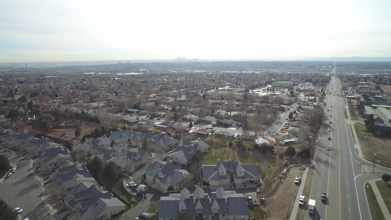 Aerial view over a suburban road with cars, houses, and a pale sky.