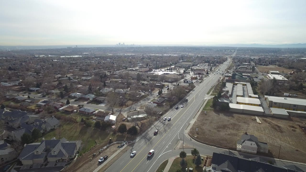 Aerial view of a suburban road with homes, low buildings, cars, and cloudy sky.