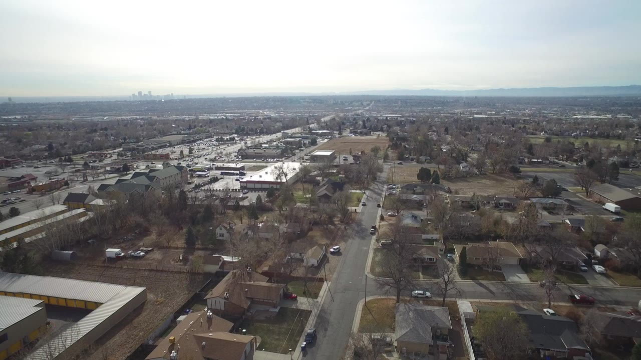 Aerial view over suburban streets, homes, trees, and distant skyline.