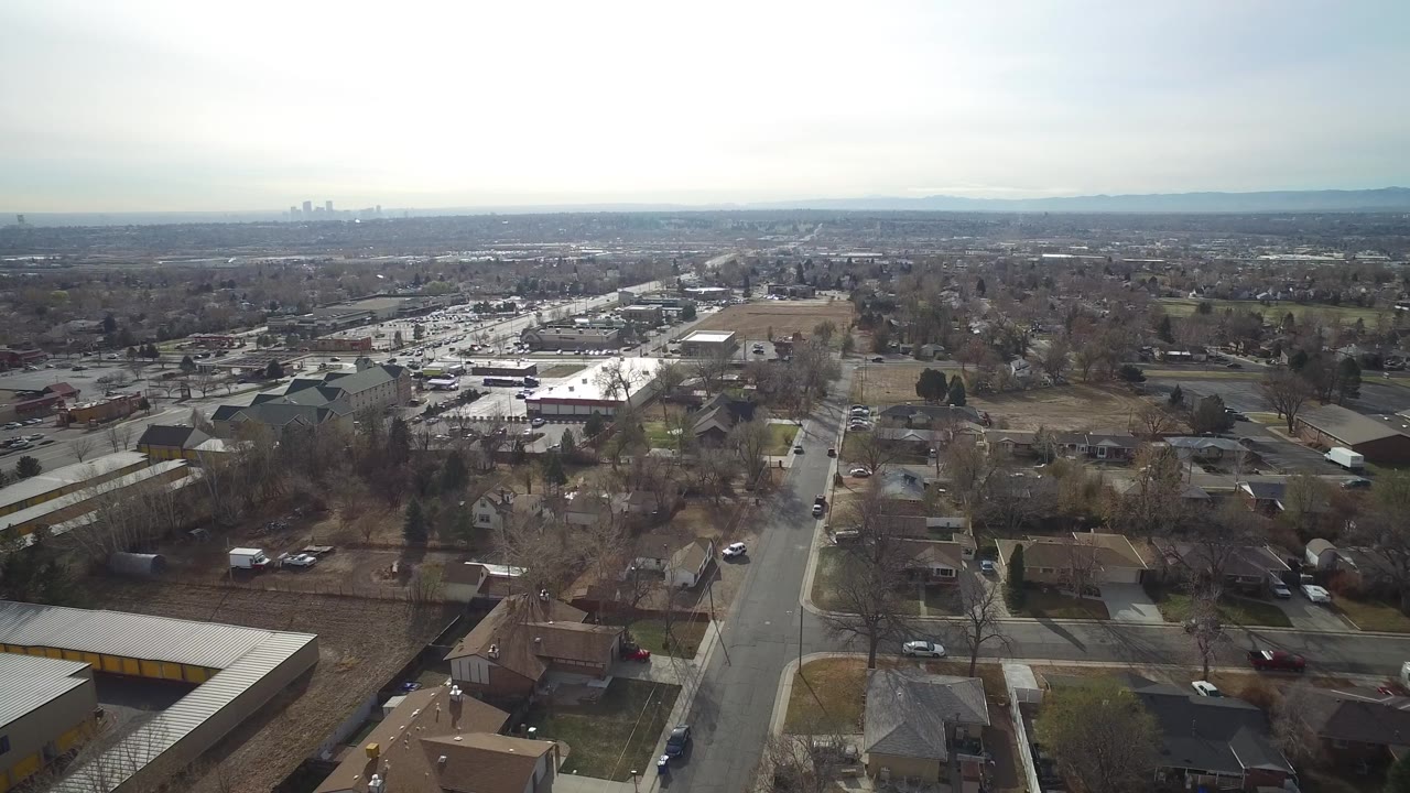 Aerial view across Westminster, Colorado, showing neighborhood blocks, a road running toward the horizon, open ground, distant hills, and no visible people.