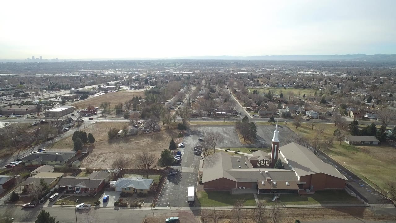 Aerial view over a suburban church-like building, streets, homes, and distant skyline.