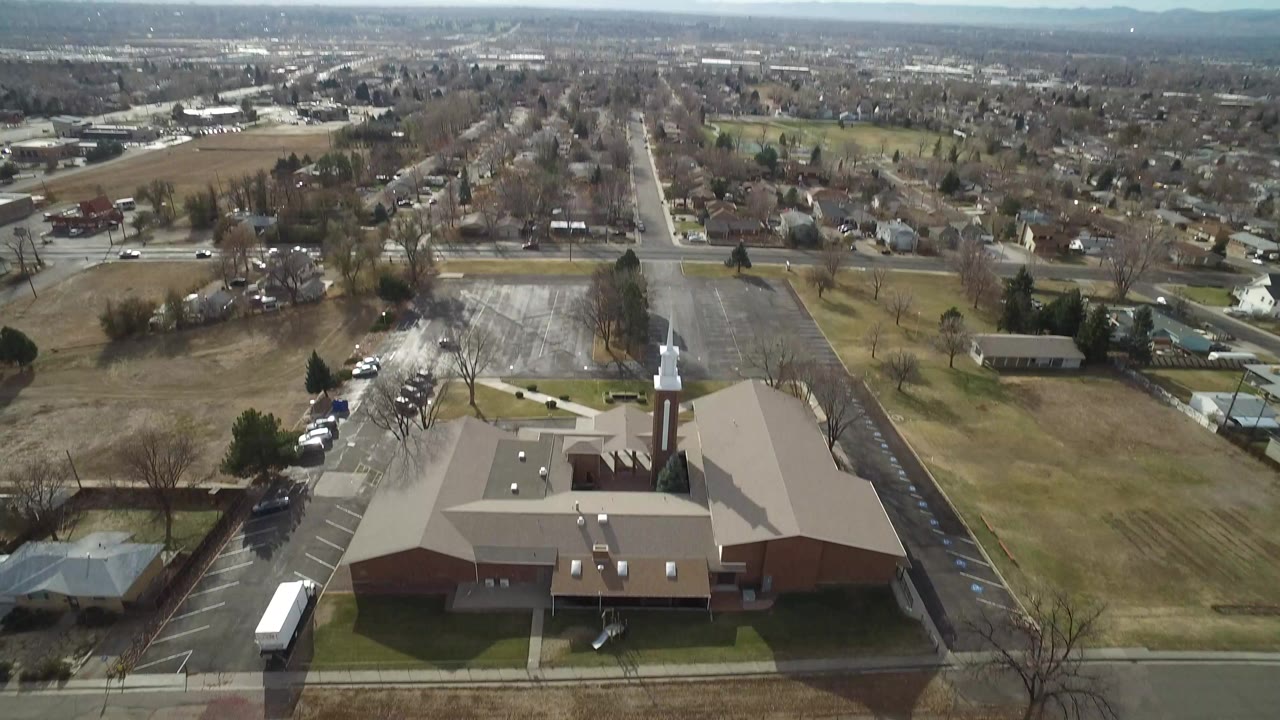 Aerial view of a tan-roofed brick building, parking lot, streets, and open land.
