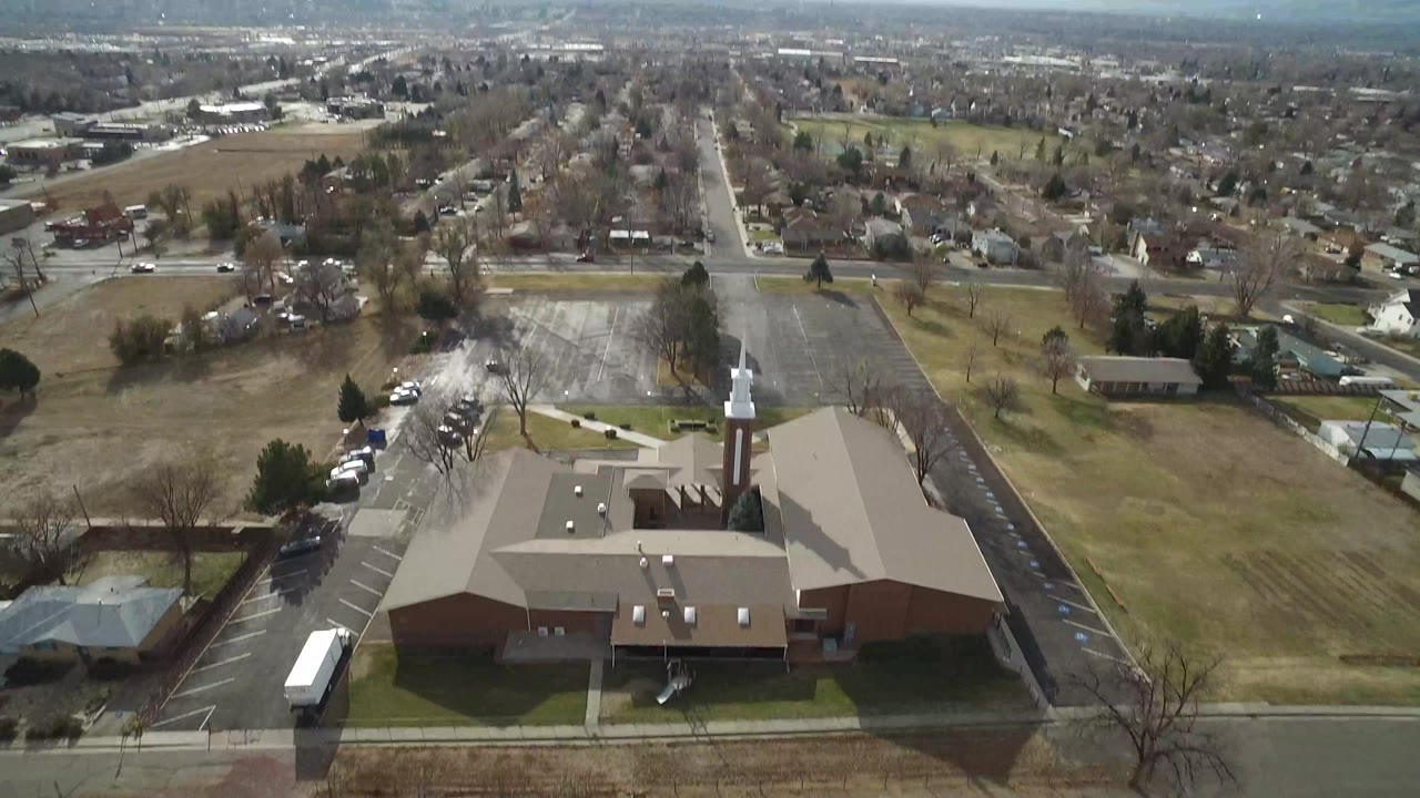 Aerial view in Westminster, Colorado with the steepled brick building centered lower in frame, a road across the foreground, open lots to the right, parked cars nearby, and no visible people.