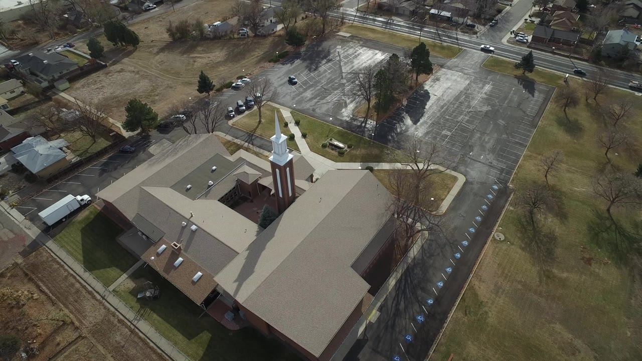A high aerial frame over Westminster, Colorado shows a brick church with a white steeple, tan roofs, adjacent parking lot, roads, and winter trees, with no visible people.