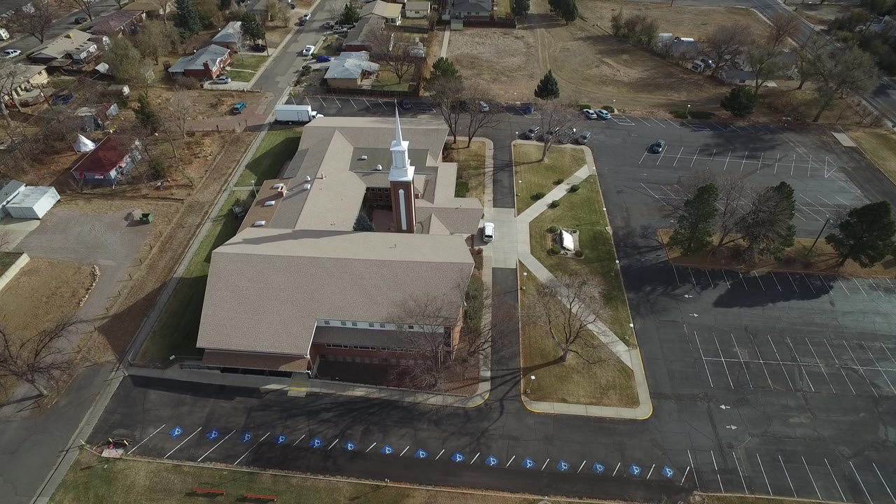 Aerial view in Westminster, Colorado showing a tan-roofed church with a white steeple, surrounding parking lots, sidewalks, nearby houses, and no visible people.