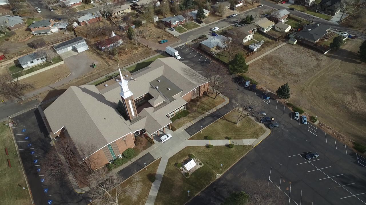 High aerial frame over a brick church sanctuary in Westminster, Colorado, showing the white steeple, empty parking rows, sidewalks, winter-brown grass, nearby houses, and no visible people.