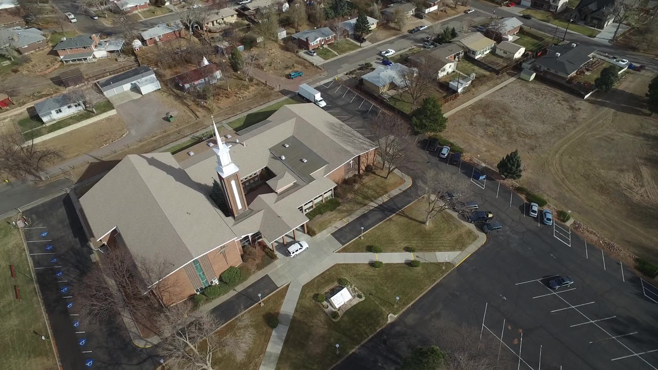 Second aerial frame in Westminster, Colorado, with the church centered lower in view, the parking lot along the right edge, residential homes in the background, and no visible people.
