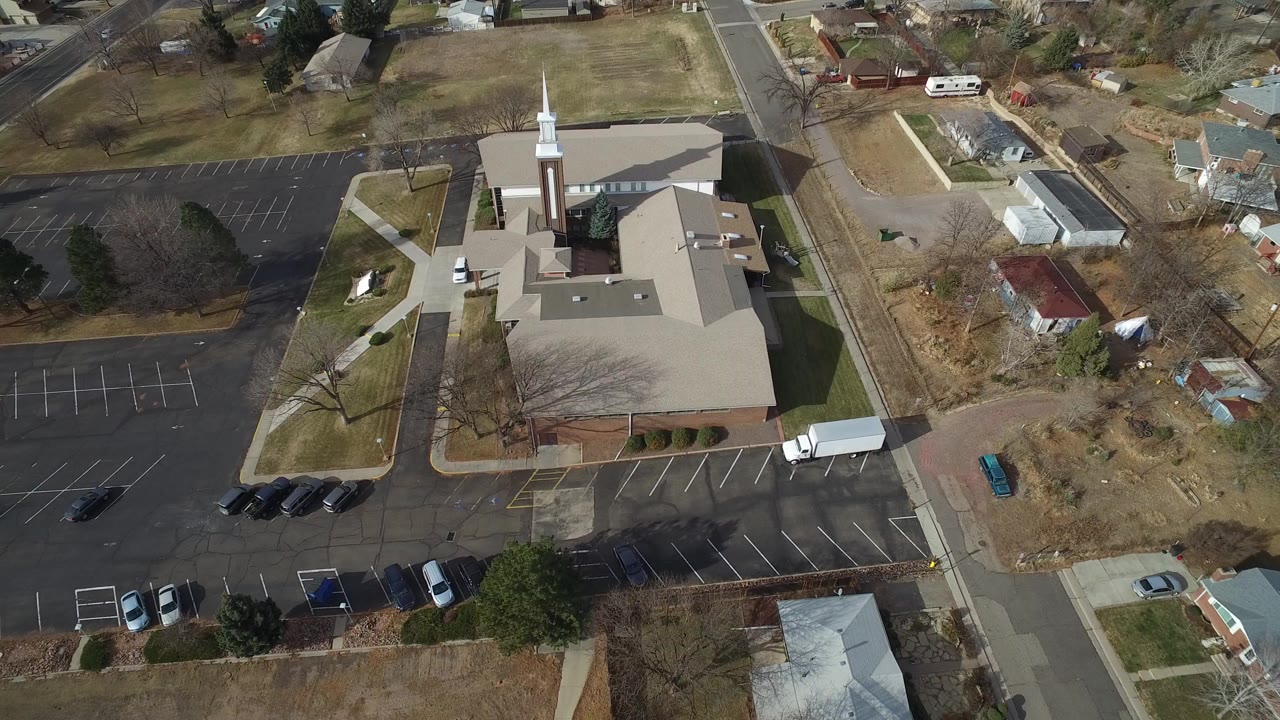 Overhead frame in Westminster, Colorado showing a light-roofed steepled building, dark parking lots, adjacent streets, parked cars, leafless trees, and no visible people.